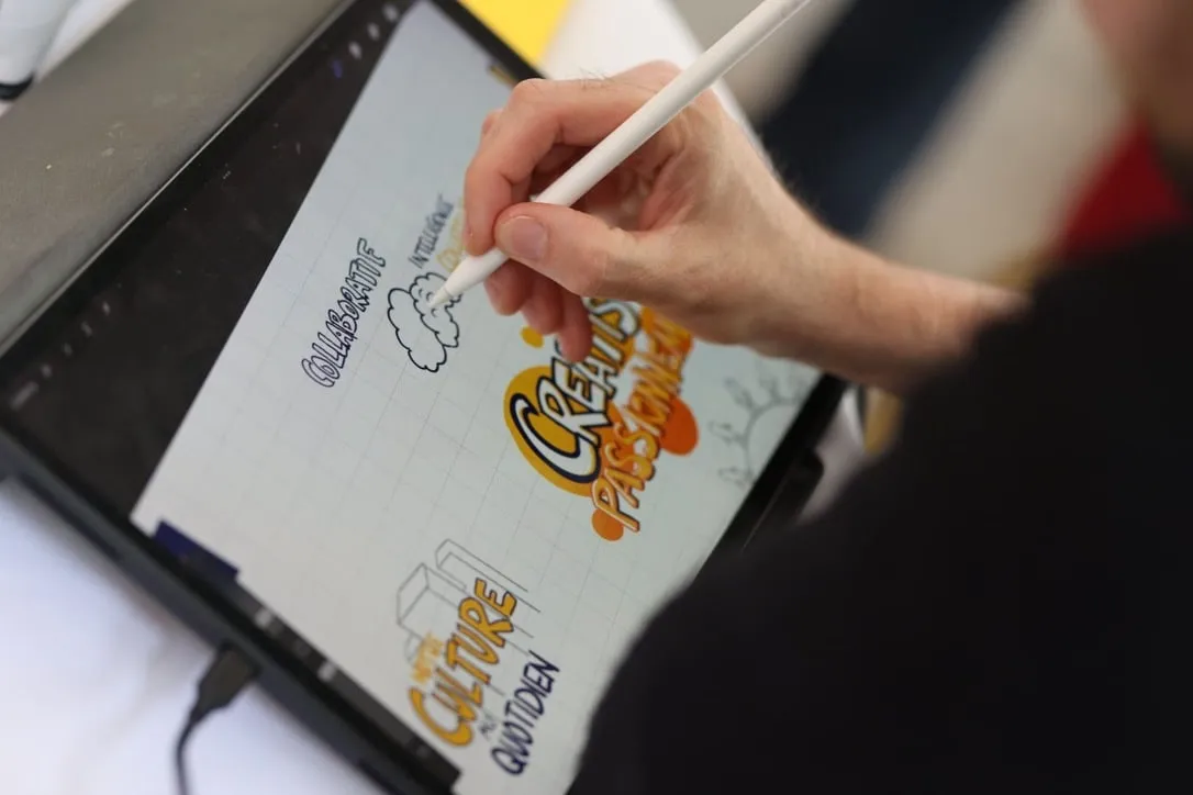 A hand draws on a tablet with a stylus during a session, lettering words and small sketches in orange, yellow and black.