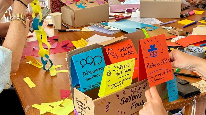 Hands at a session table build cardboard prototype boxes, with hand-lettered paper cards, a paper chain and marker pens.