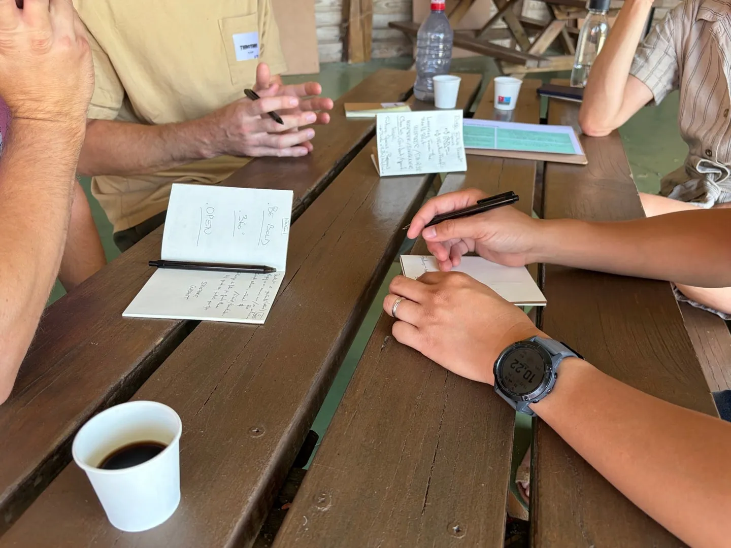 Hands writing in notebooks on a wooden table during a two-day workshop, a coffee cup in the foreground.